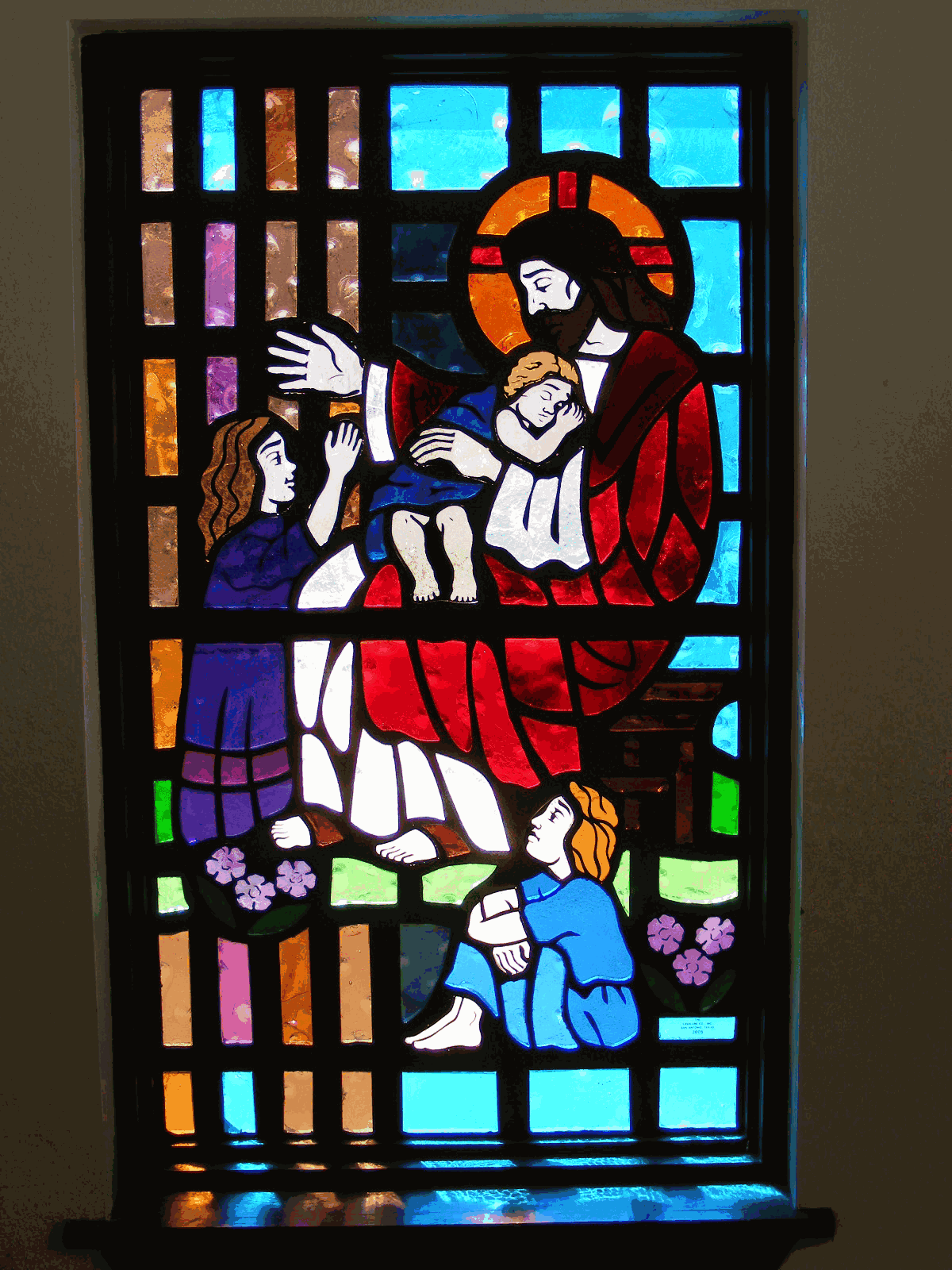 Stained glass window depicting a religious figure holding a child with two other figures nearby, vivid blue, red, and gold tones
