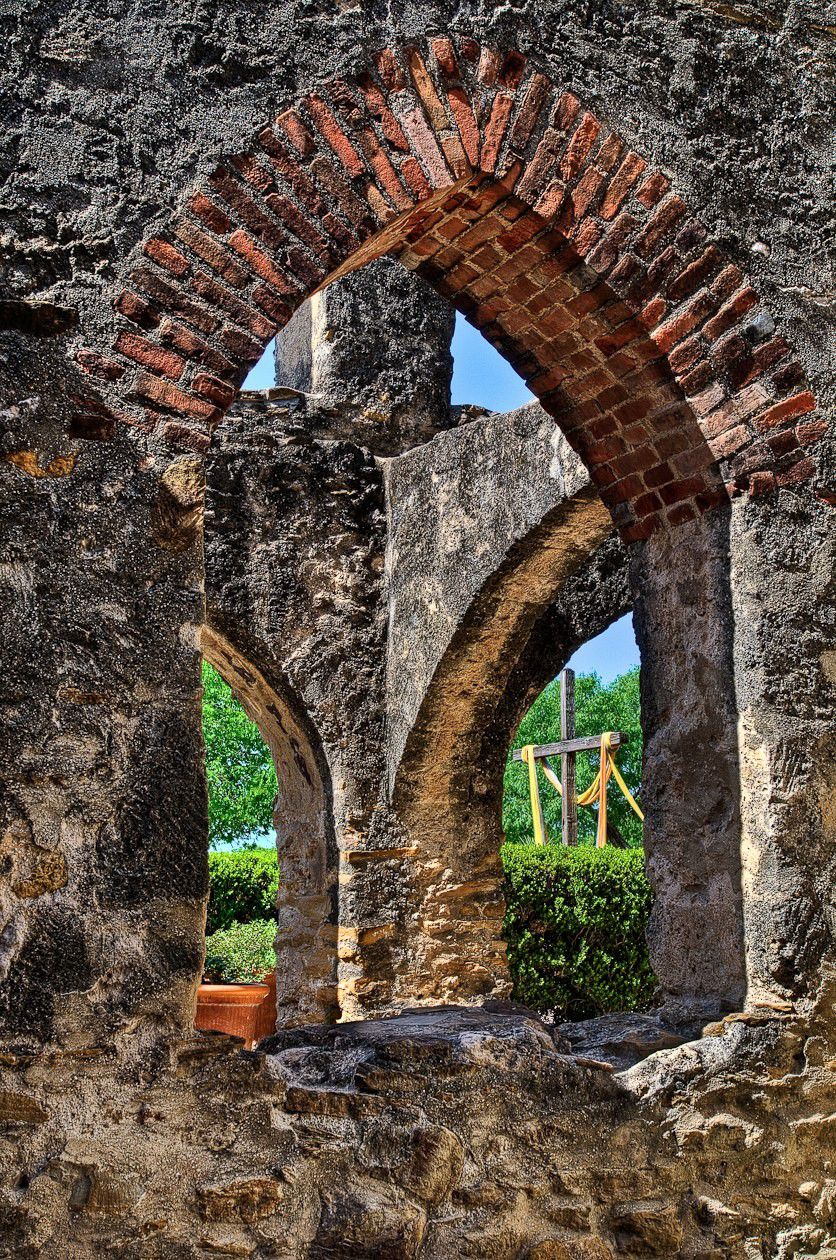 Stone archway ruins with brick arch framing a green garden and wooden cross outside, warm earthy tones