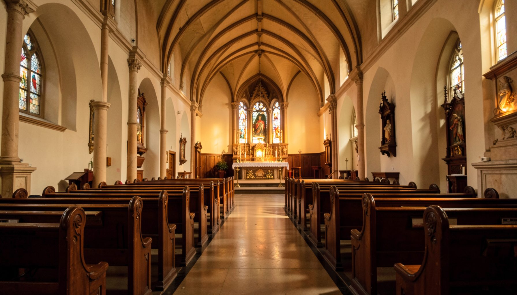 Warm interior of a Catholic church with wooden pews, soft amber lighting, and a distant altar beneath an arched ceiling