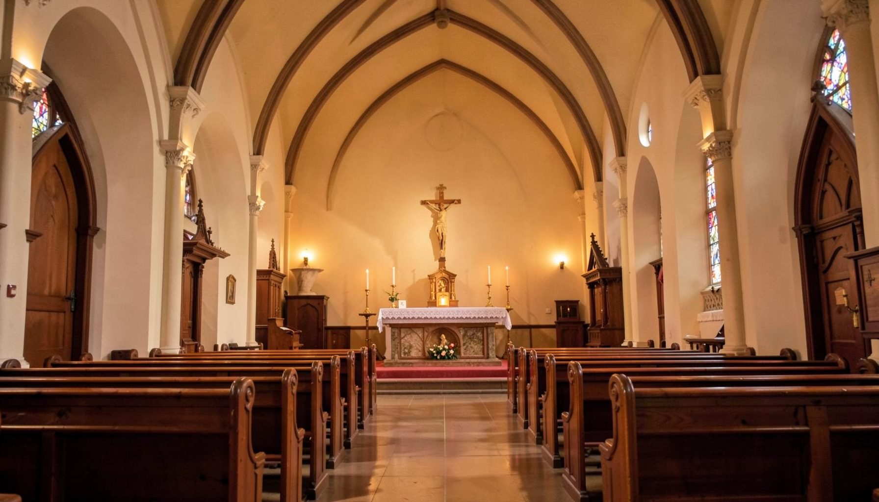 Warm interior of a Catholic church with wooden pews, soft amber lighting, and a simple altar beneath a high arched ceiling